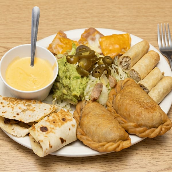 A platter of Mexican appetizers including quesadillas, taquitos, empanadas, guacamole, jalapeños, and cheese dip.