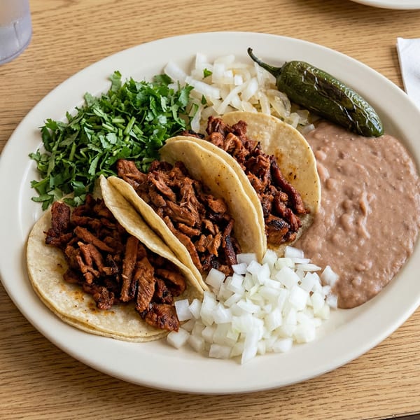 Three tacos al pastor with cilantro, onions, refried beans, and a grilled jalapeño on a white plate.