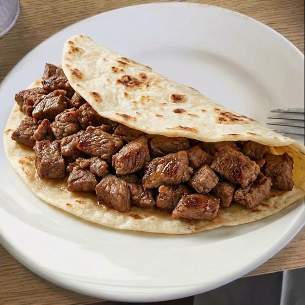 A close-up of a flour tortilla filled with generous chunks of seasoned steak on a white plate.