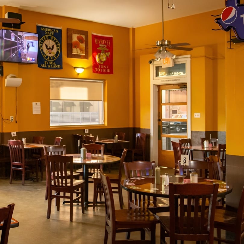 Empty restaurant dining room with round tables, wooden chairs, and military flags on orange walls.