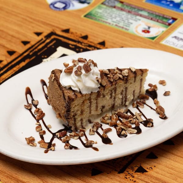 Slice of cake with whipped cream, pecans, and chocolate drizzle on a white plate.