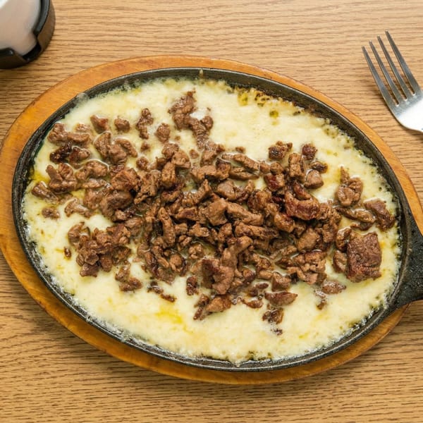 Sizzling skillet of melted white cheese topped with browned diced meat, on a wooden table.